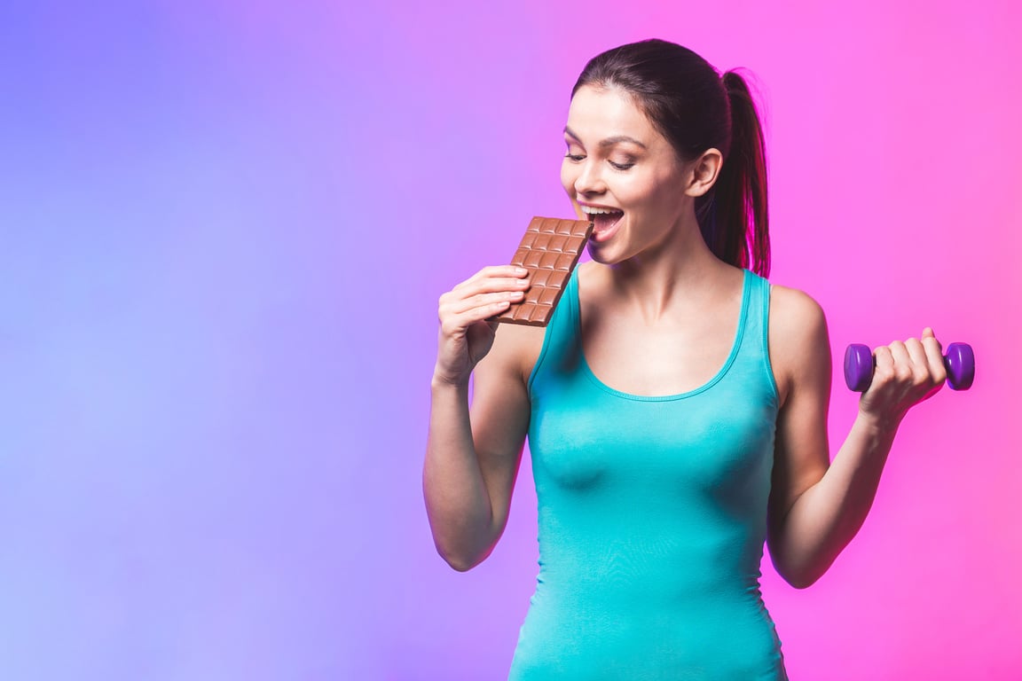 Chocolate against dumbbells! Portrait of young beautiful woman making choice between sport and unhealthy food isolated against white background.