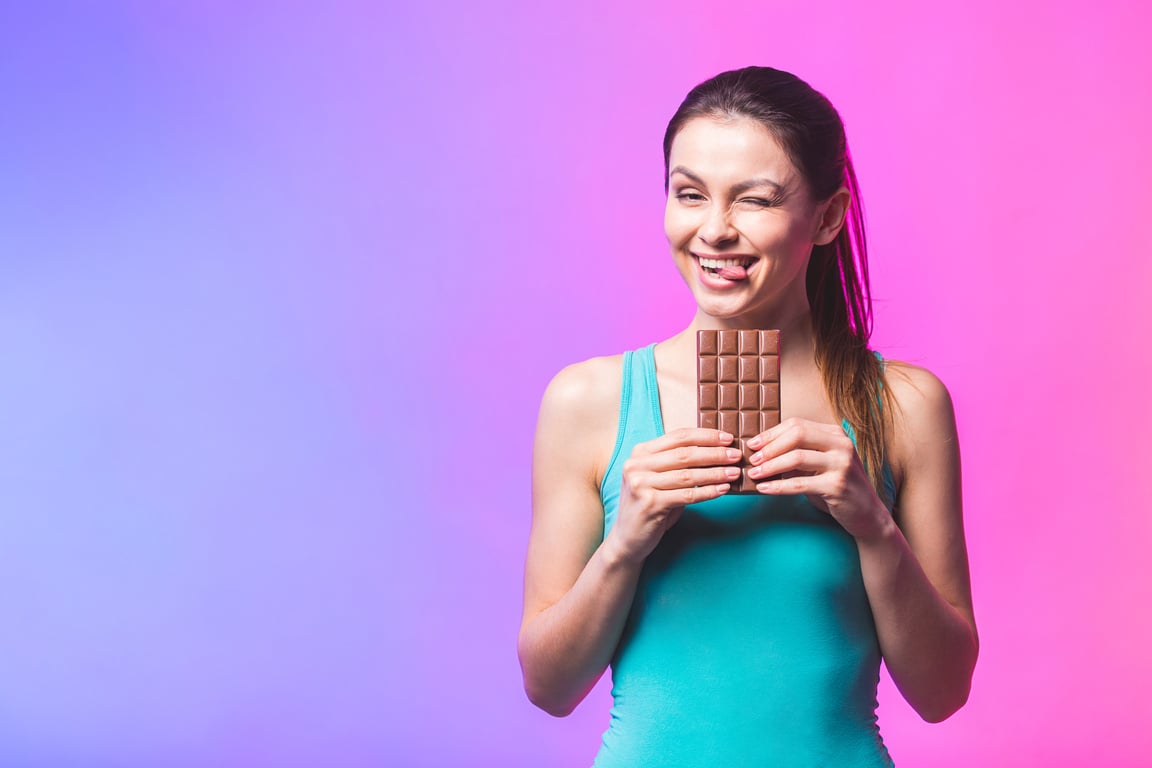 Happy young fitness beautiful lady eating chocolate and smiling. Girl tasting sweet chocolate. Young woman with natural make up having fun and eating chocolate isolated against white background.